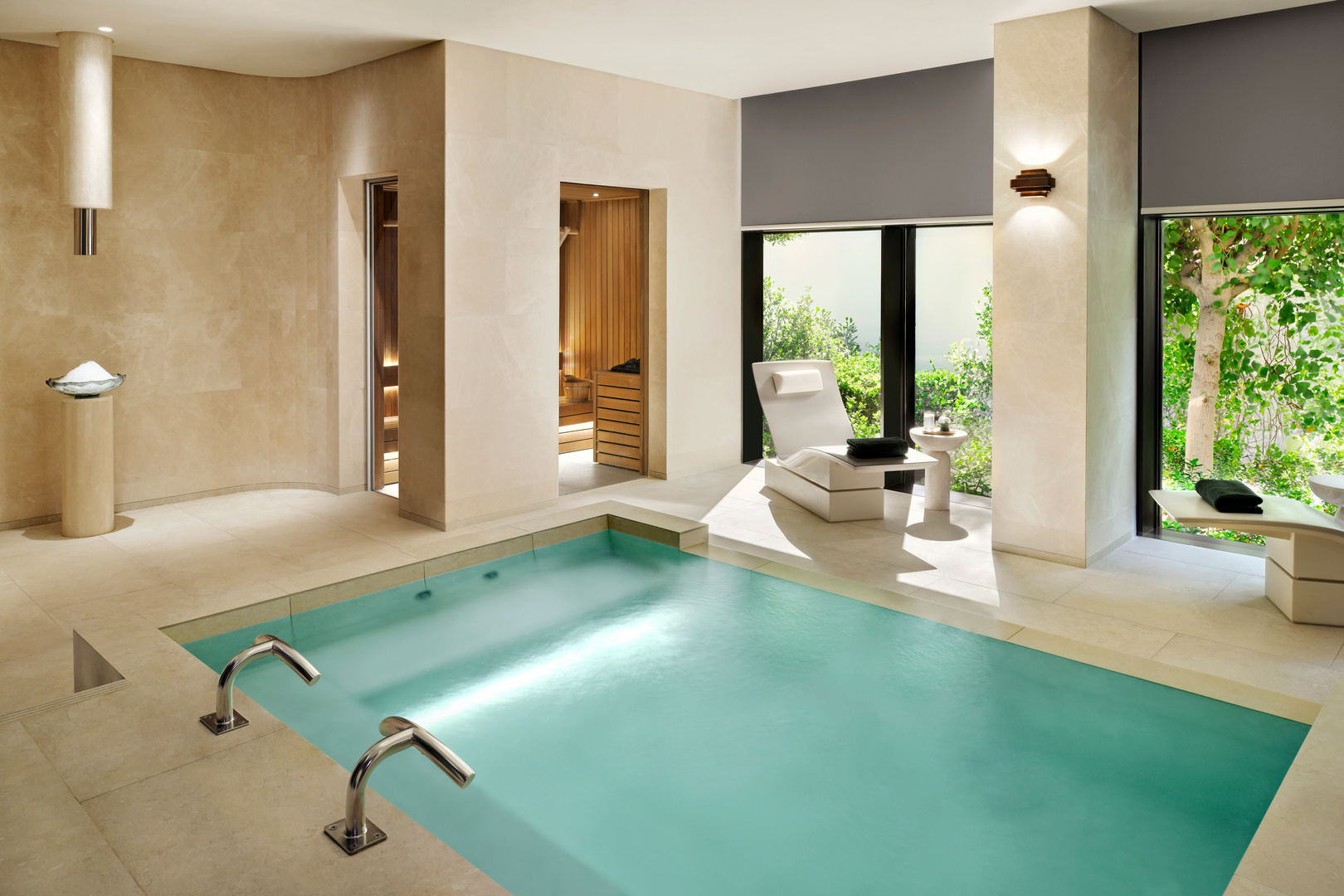 Luxurious indoor spa suite featuring a private heated pool, modern lounge chairs, a sauna with wooden interior, and large windows overlooking lush greenery. The space is designed in calming neutral tones, creating a serene and tranquil wellness retreat.